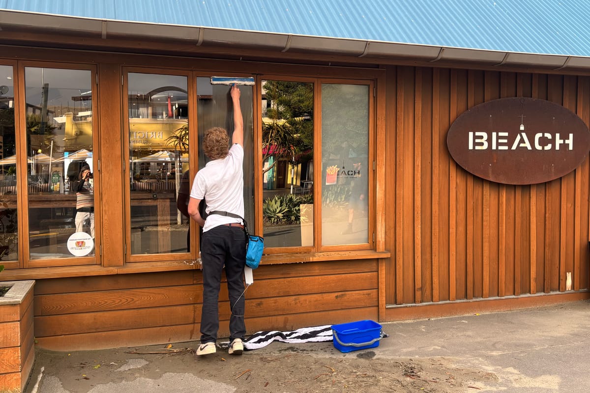 Luca cleaning the windows of a cedar-clad shopfront from the ground, drop cloth down