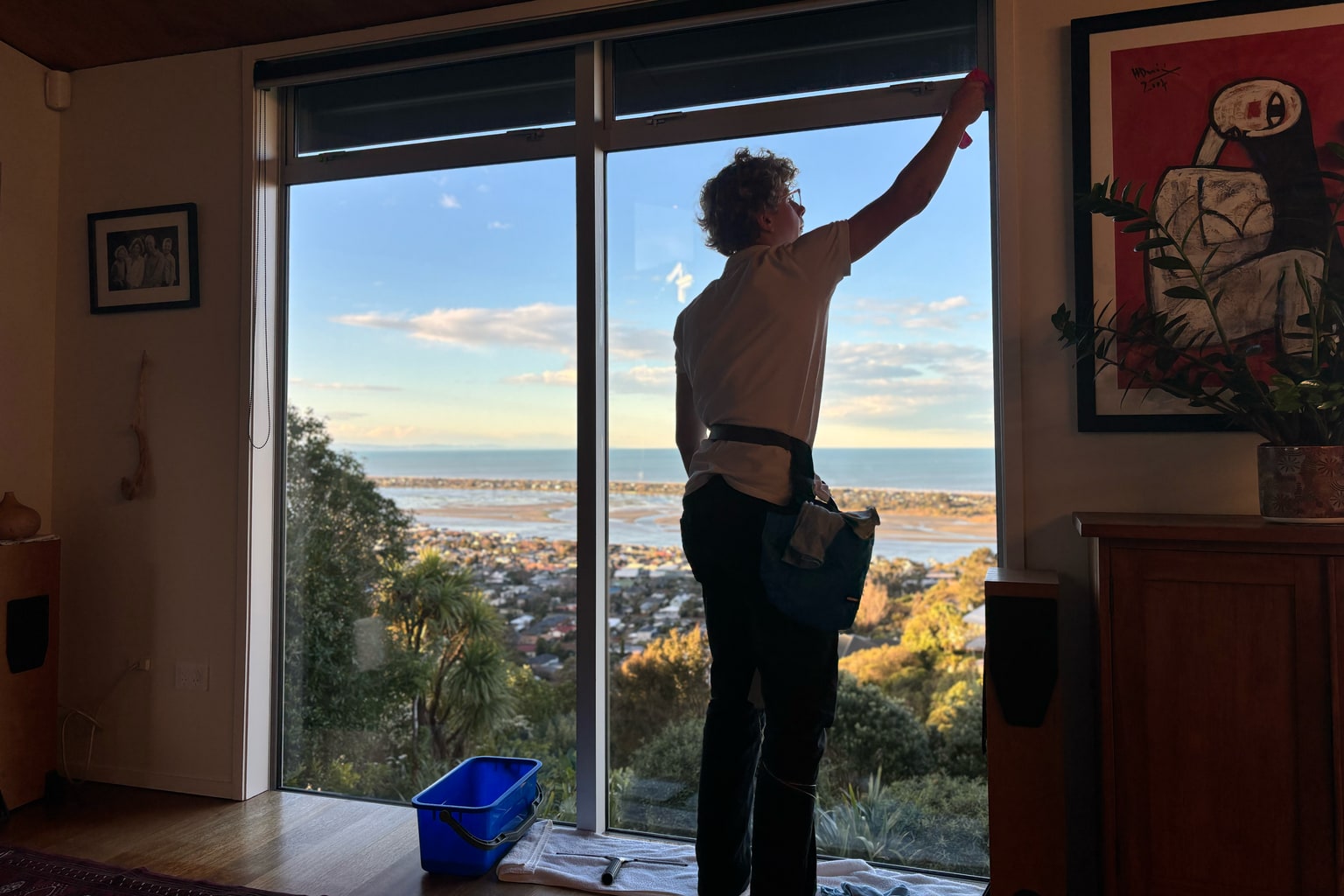 Luca cleaning the inside of a full-height window above the Christchurch estuary